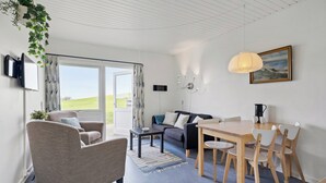 A modern living room with a sofa, armchair, dining table, and chairs. There is a television mounted on the wall, a painting above the table, and a window with curtains. - Cozy Brenderup Apartment on the Beach (Middelfart)