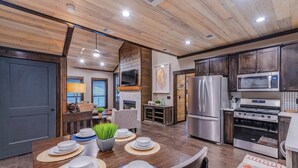 A modern kitchen with wooden ceiling, stainless steel appliances, and a dining area with a wooden table set for four. - Glorious Sunset - Hot Tub - Fire Pit - Game Room - Forest Views - Sleeps 8 (BROKEN BOW)