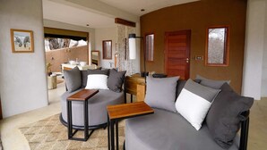 Ein modernes Wohnzimmer mit einem grauen Sofa, zwei Sesseln, einem Holztisch und einem kleinen Beistelltisch. Eine Holztür und ein Fenster mit Blick auf Bäume sind ebenfalls zu sehen.