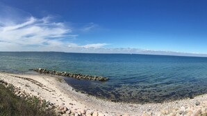 Un paysage côtier offrant une vue dégagée sur la mer, une plage de sable et une zone herbeuse avec un sentier menant à un bâtiment blanc.
