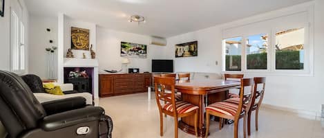 A spacious living room with a black leather sofa, a dining table with six chairs, and a kitchen area with wooden cabinets and a TV.