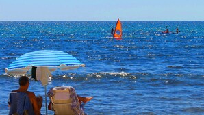 Eine Strandansicht mit zwei Personen, die unter einem gestreiften Sonnenschirm sitzen, einem Segelboot auf dem Wasser und einem Sandstrand.