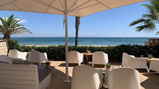 Eine Terrasse am Strand mit einem großen Sonnenschirm, weißen Stühlen und Blick aufs Meer.