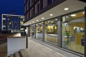 L’extérieur d’un hôtel moderne, avec un comptoir d’accueil, des portes vitrées et un espace salon visible à travers les fenêtres.