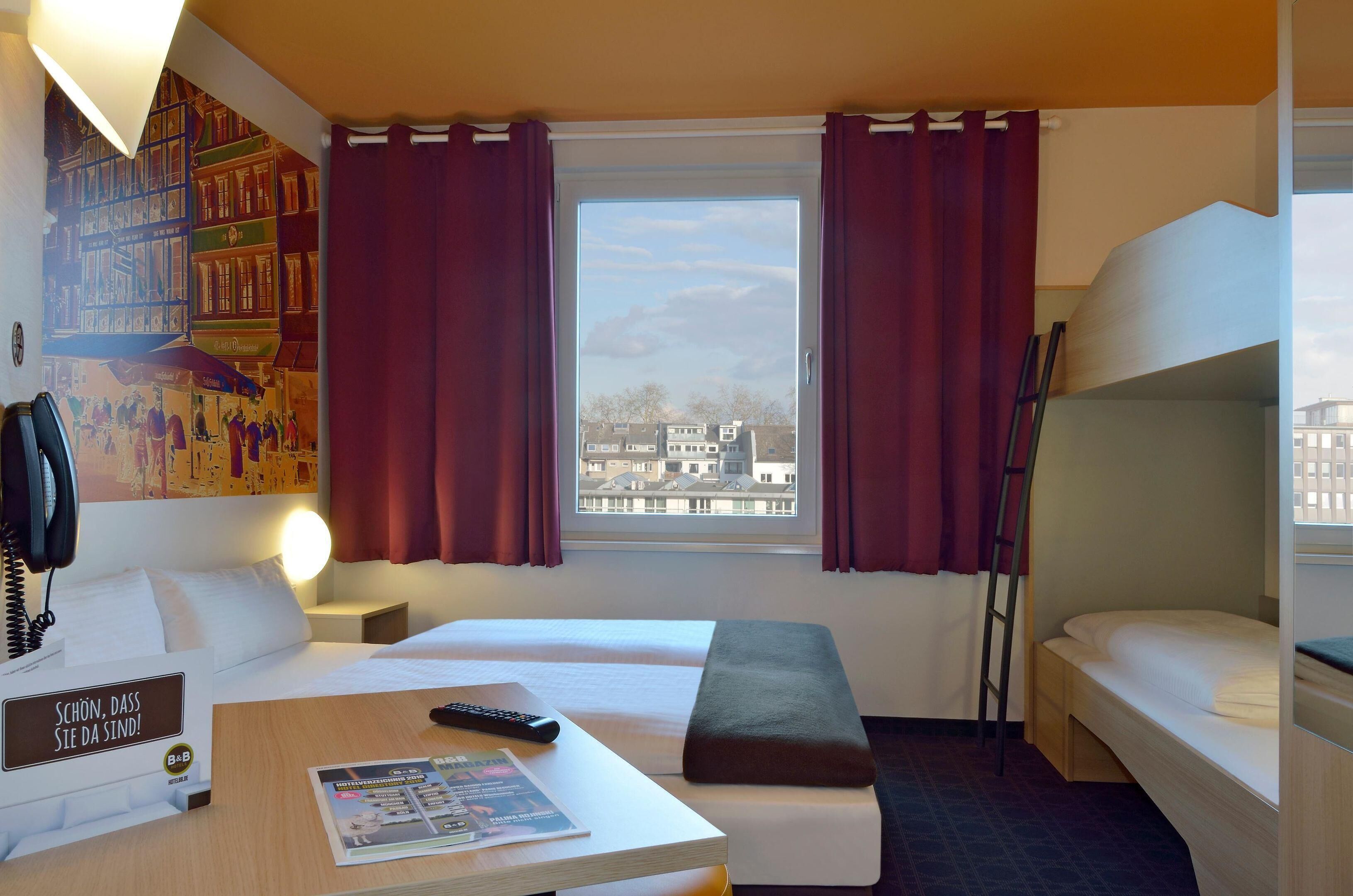 A hotel room with two beds, a desk with a remote control, a lamp, and a window with curtains.