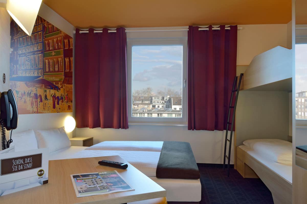 Une chambre d’hôtel avec deux lits, un bureau équipé d’une télécommande, une lampe et une vue sur les immeubles par la fenêtre.