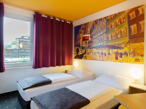 Een hotelkamer met twee bedden, een groot raam met gordijnen, een schilderij van een straatbeeld en een nachtkastje met een lamp.