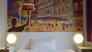 Une chambre d’hôtel avec un lit, deux lampes de chevet et une grande fresque murale représentant une scène animée de rue.
