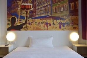 Une chambre d’hôtel avec un lit, deux lampes de chevet et une grande fresque murale représentant une scène animée de rue.