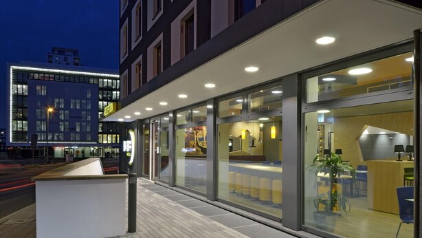 A modern hotel exterior with a reception desk, glass doors, and a seating area visible through the windows.