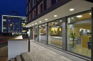 Een modern hotelgebouw met een receptiebalie, glazen deuren en een zithoek die door de ramen zichtbaar is.