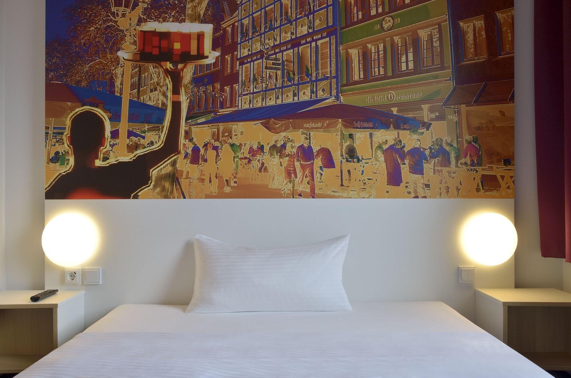 A hotel room with a large bed, two bedside lamps, and a wall mural depicting a busy street scene.