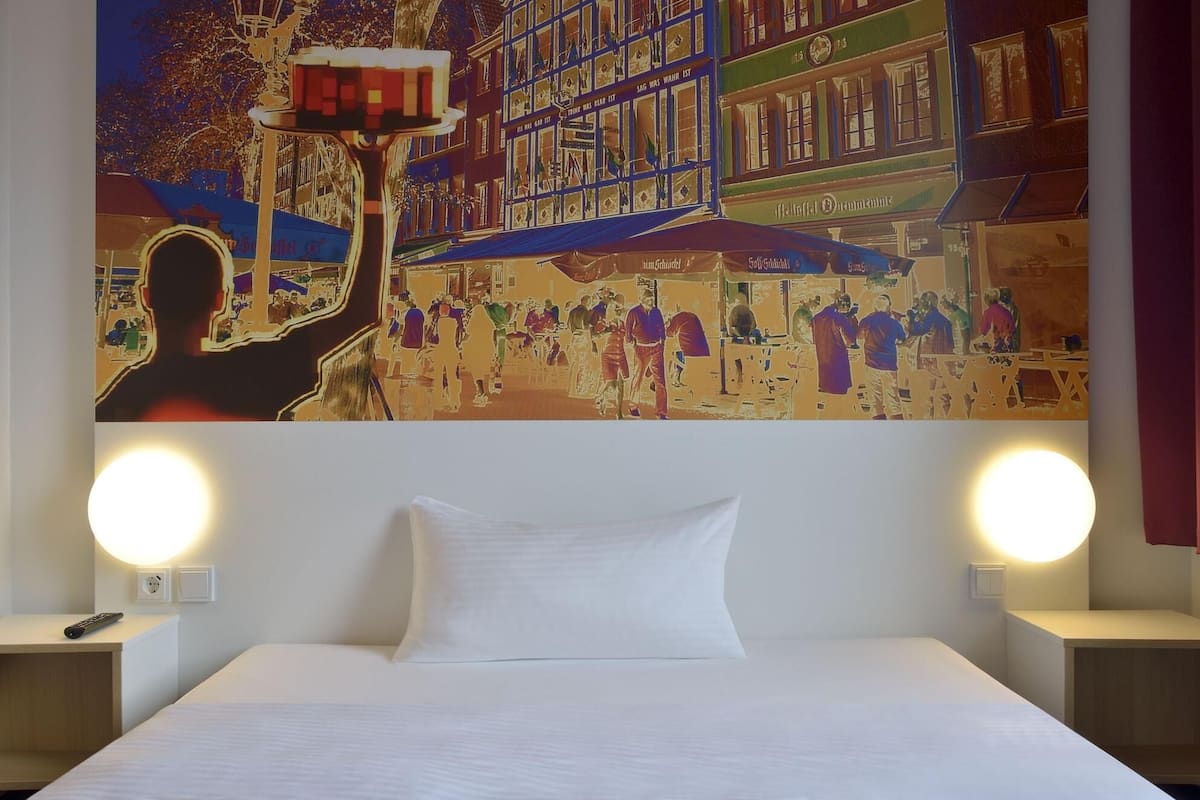 Une chambre d’hôtel avec un lit, deux lampes de chevet et une grande fresque murale représentant une scène animée de rue.