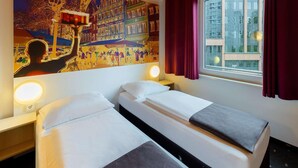 Une chambre d’hôtel avec deux lits, une grande fresque murale, une fenêtre avec des rideaux et une vue sur les bâtiments environnants.