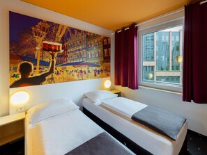 Hotelkamer met twee bedden, een groot muurschilderij, een raam met gordijnen en uitzicht op gebouwen.