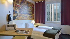 A hotel room with a bed, a desk with a phone and a magazine, a window with curtains, and a wall with a cityscape mural.