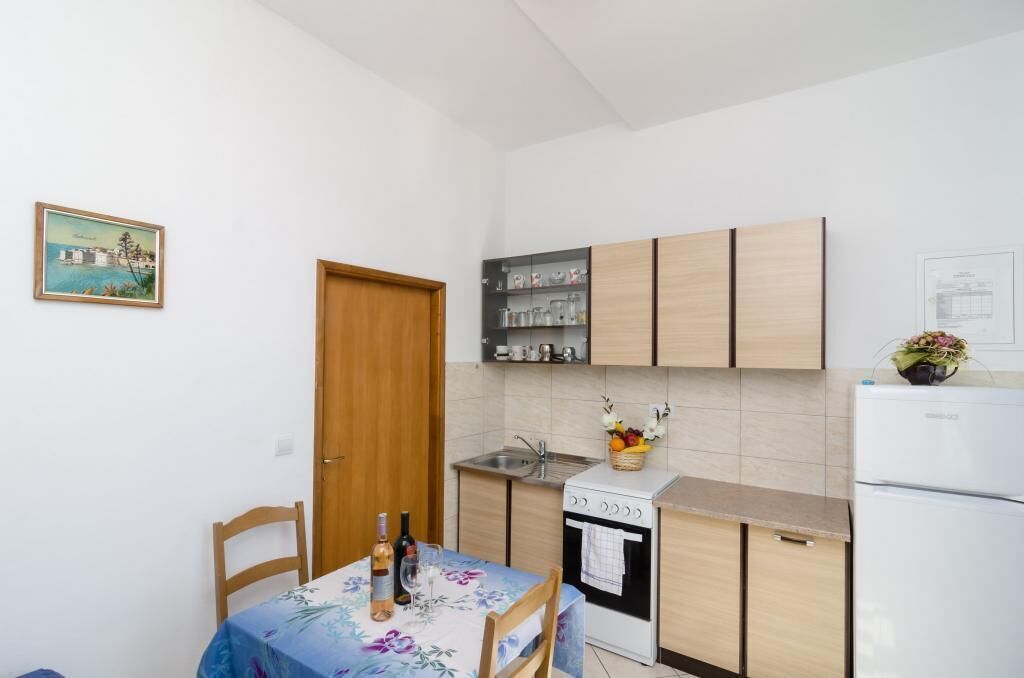 A compact kitchen with a wooden door, a stove, a sink, and a refrigerator. There is a table with chairs and a vase of flowers.