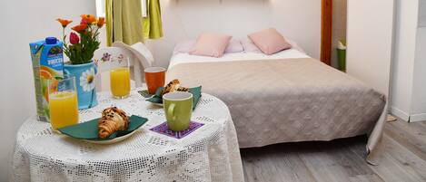 A neatly arranged breakfast table with croissants, juice, and a vase of flowers. A bed with a pink pillow and a wall-mounted shelf with decorative items are also visible.