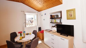 Ein gemütliches Zimmer mit Holzbalkendecke, einem Esstisch mit Stühlen, einem Fernseher auf einer Holzkommode und einem Fenster mit Blick auf Gebäude.