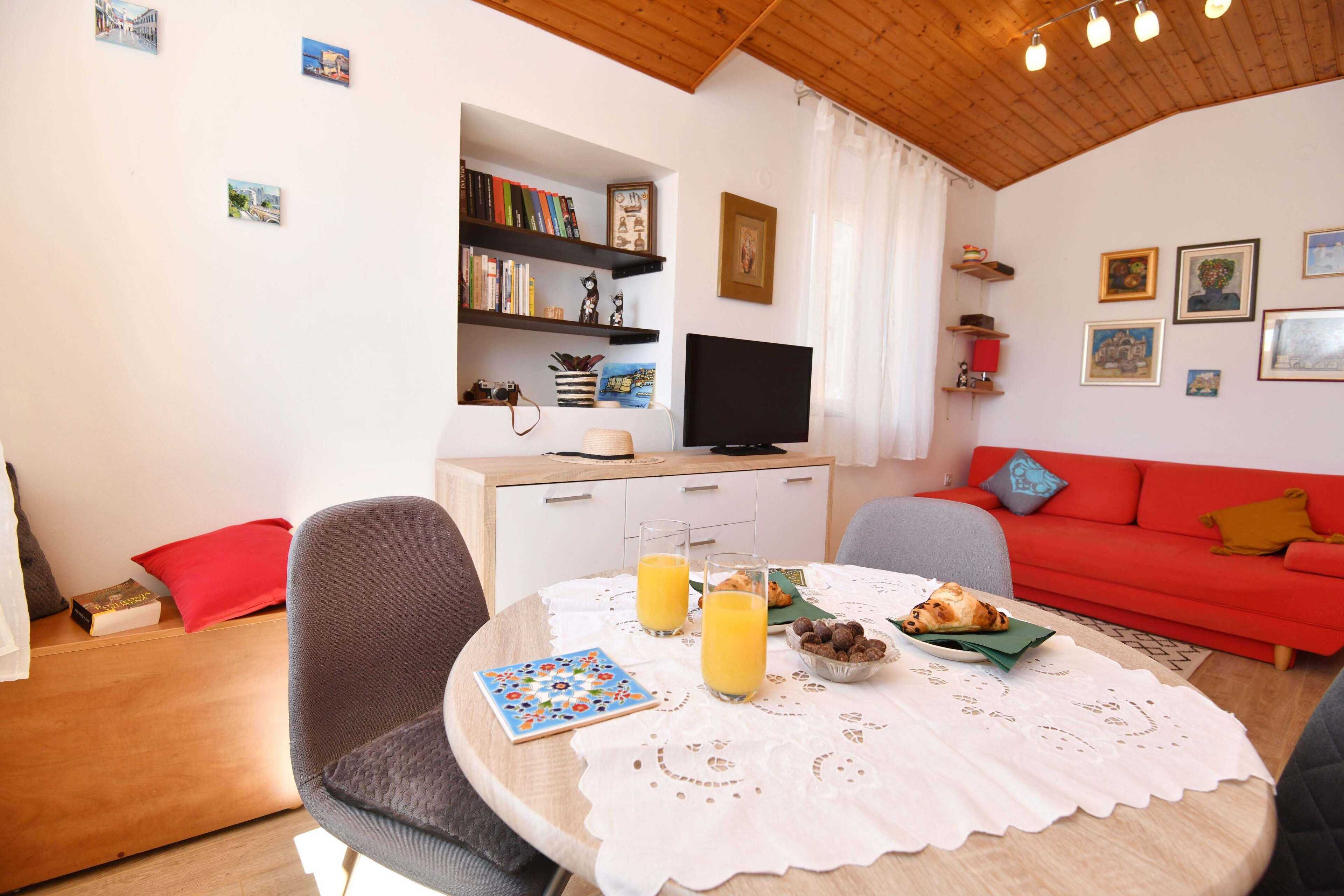 A cozy living room with a round table, chairs, a red sofa, a bookshelf, and a television.