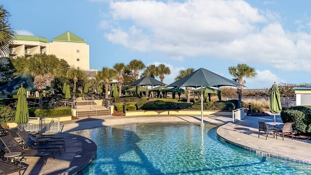 A pool area with lounge chairs, umbrellas, and a building in the background. - Hilton Head | Island Beach Stay | Pool & Beach | 2BR (Hilton Head)
