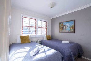 A room with two beds, a window with red blinds, and a painting on the wall.