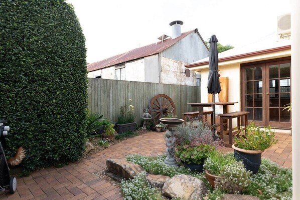 A rustic outdoor patio with a wooden table and chairs, a stone fountain, and a wooden wheel.