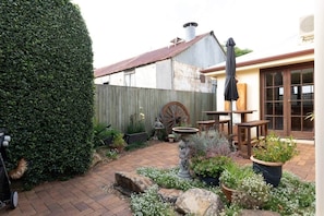 A rustic outdoor patio with a wooden table and chairs, a stone fountain, and a wooden wheel.