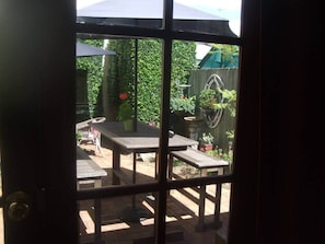 Blick durch eine Glastür auf einen Außenbereich mit einer Holzterrasse, einem Holztisch und Bänken, umgeben von Topfpflanzen und einem Holzzaun.
