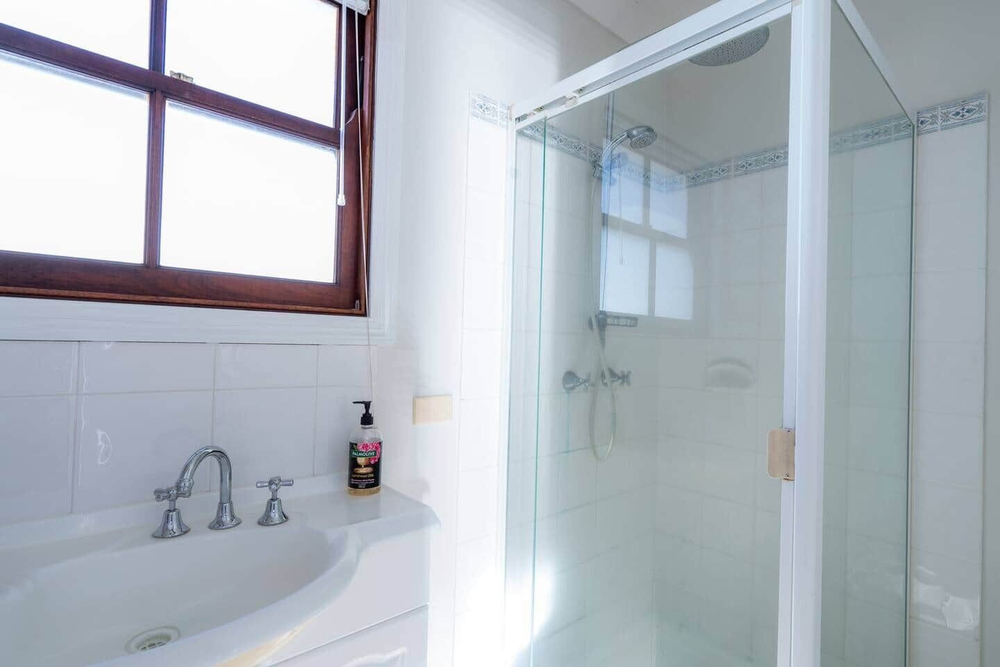A bathroom with a glass-enclosed shower, a white sink with two faucets, and a window above the sink.
