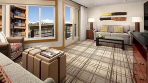 A modern hotel room with a large window offering a mountain view, a cozy seating area with a sofa and armchairs, a small table with a lamp, and a flat-screen TV.