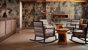A modern lounge area with a stone wall, wooden ceiling, and a fireplace. There are four patterned chairs and a round wooden table.