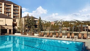 A swimming pool with lounge chairs, a mountain backdrop, and a multi-story building.