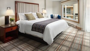 A hotel room with a large bed, two bedside tables with lamps, a view of the balcony through the window, and a patterned carpet.