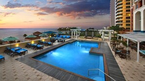 A pool area with lounge chairs and umbrellas, a building with balconies, and a view of the ocean at sunset.