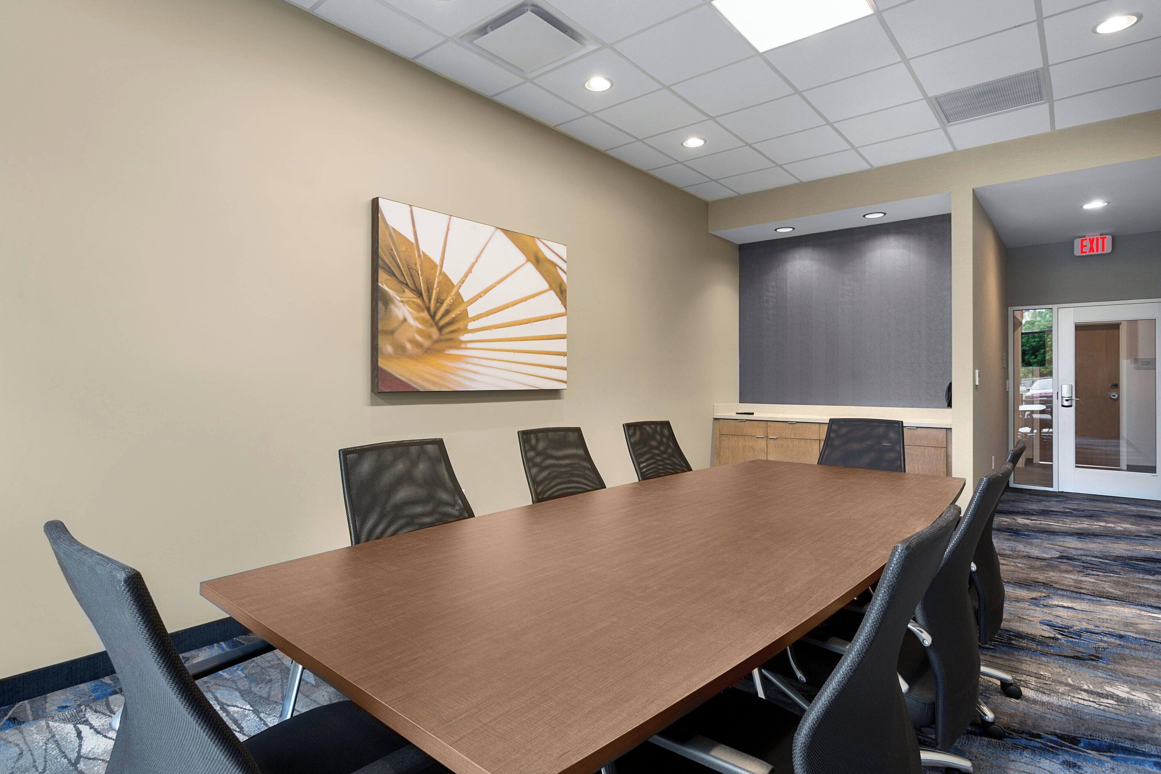 A conference room with a long table, six chairs, a projector screen, and an exit sign.