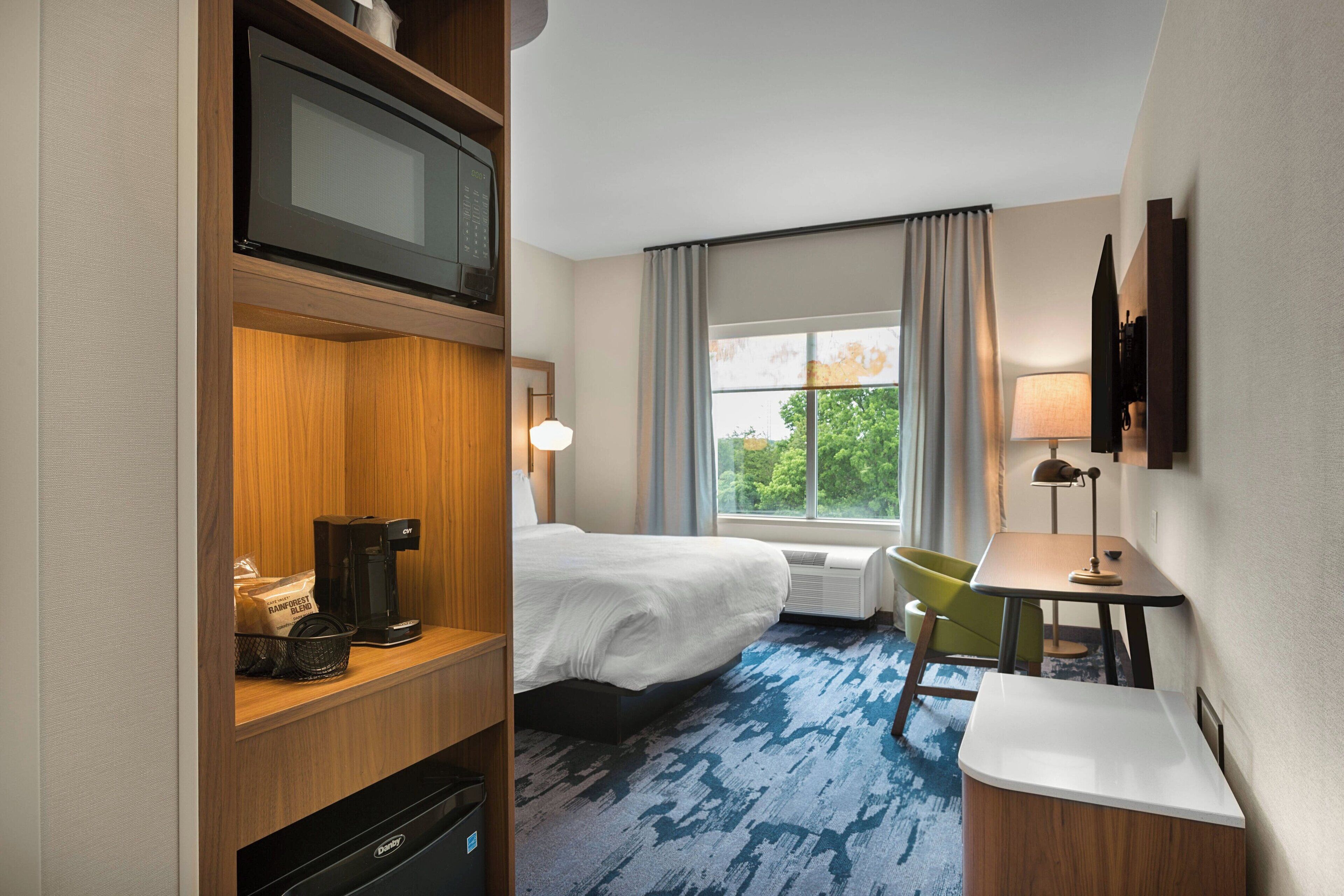 Habitación de hotel con cama, escritorio, silla, televisor, cafetera y ventana con cortinas.