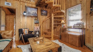 A cabin interior with a wooden interior, a fireplace, a ladder, a sofa, a coffee table, and a window with blinds.
