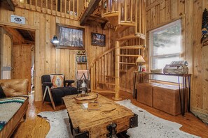 Et sommerhusinteriør med træpaneler, pejs, stige, sofa, sofabord og et vindue med persienner.