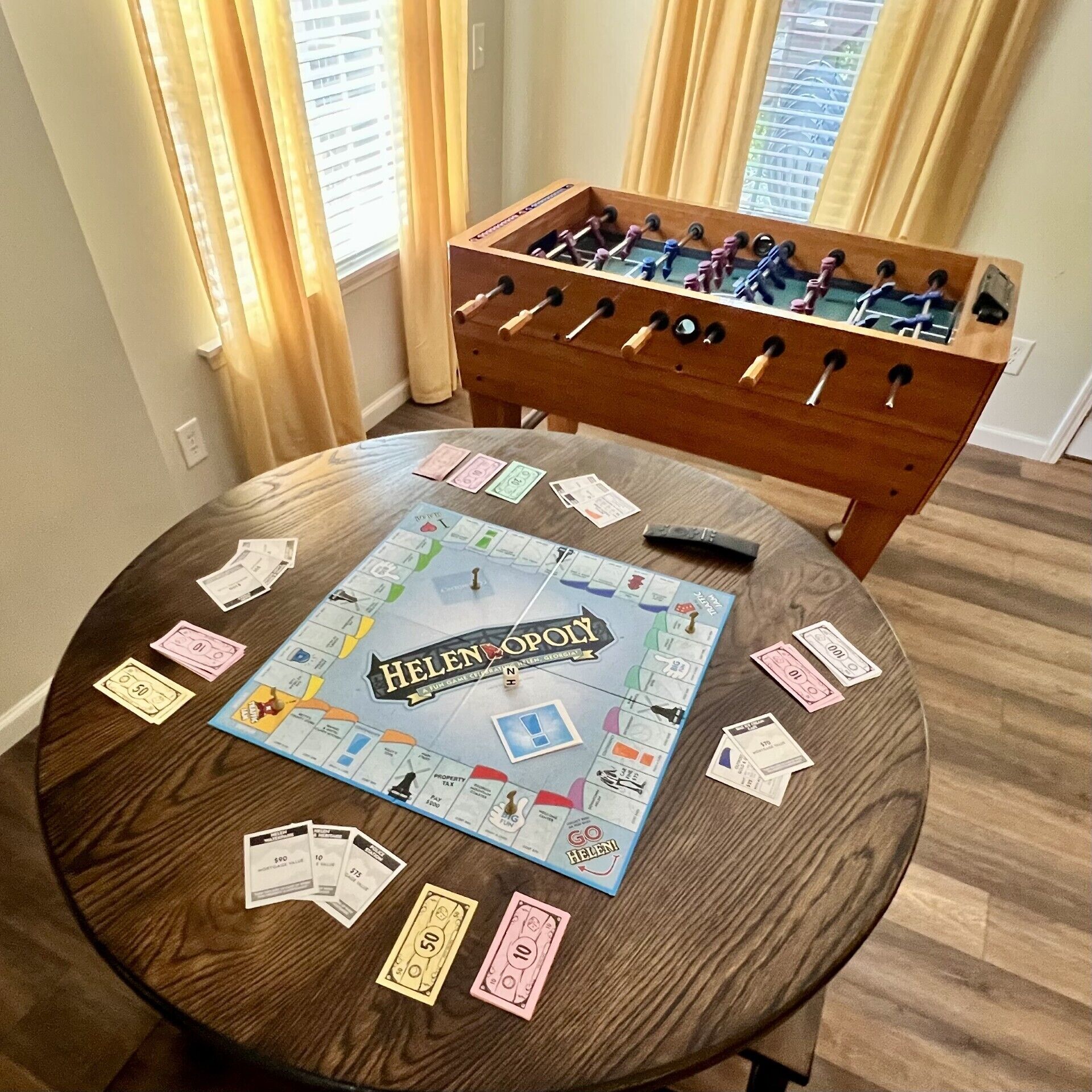 Una mesa de madera redonda con un juego de mesa llamado 'Helen's Opulence' y una mesa de futbolín al fondo.