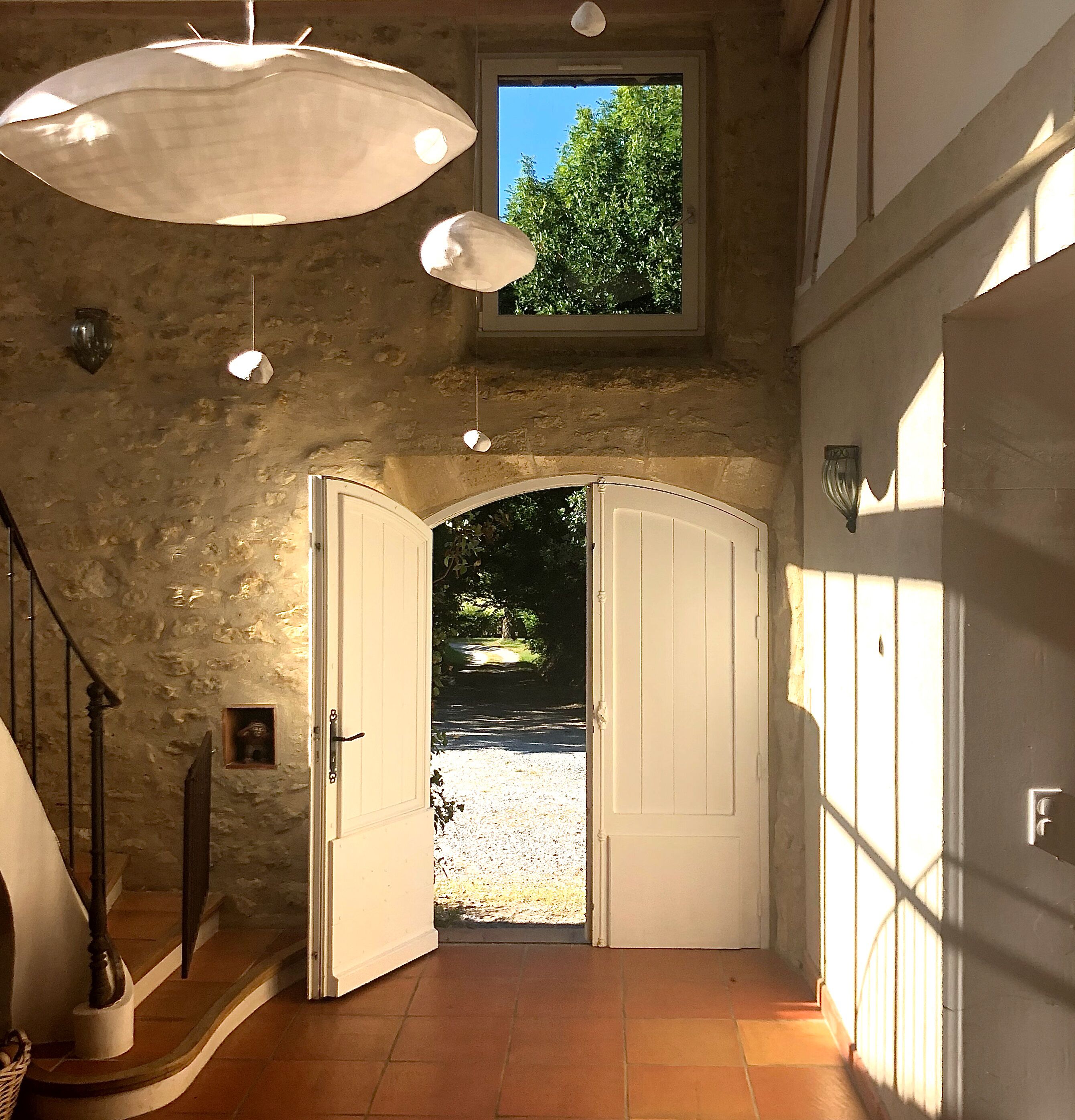 A hallway with a stone wall, a wooden staircase, and a view of a garden through open doors.