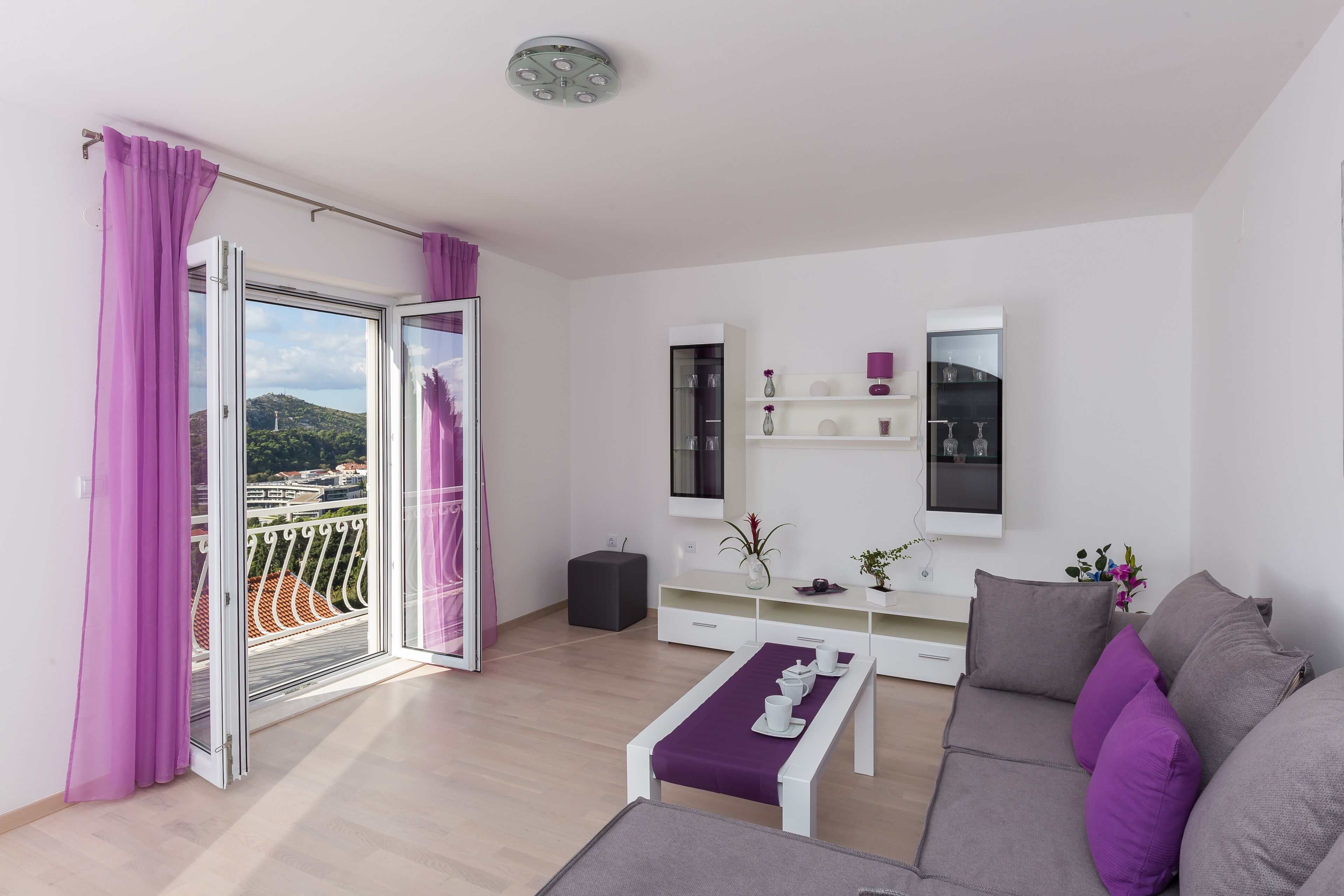 Un salon moderne avec un canapé, une table basse et un téléviseur. Il y a un balcon avec des rideaux violets et une vue sur la ville.