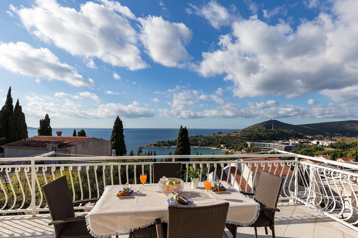 Un balcón con una mesa lista para el desayuno, con vistas a la costa, montañas y un pueblo al fondo.
