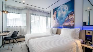 A hotel room with two beds, a desk with a chair, a large window with sheer curtains, and a wall mural of a moonlit cityscape.