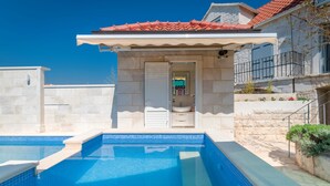 Une piscine dont le bord est revêtu de carrelage, un mur en pierre et une porte donnant accès à une salle de bain.