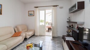 A living room with a beige sofa, a glass coffee table with fruits, a TV, and a small kitchen area with a fridge and shelves. - Apartment Alana - (Supetar)