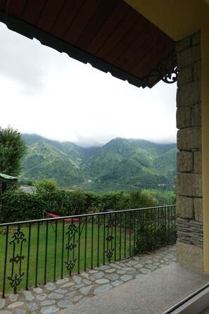 Un balcon doté d’un sol en pierre et d’une rambarde en métal, offrant une vue sur un paysage montagneux verdoyant et un ciel nuageux.