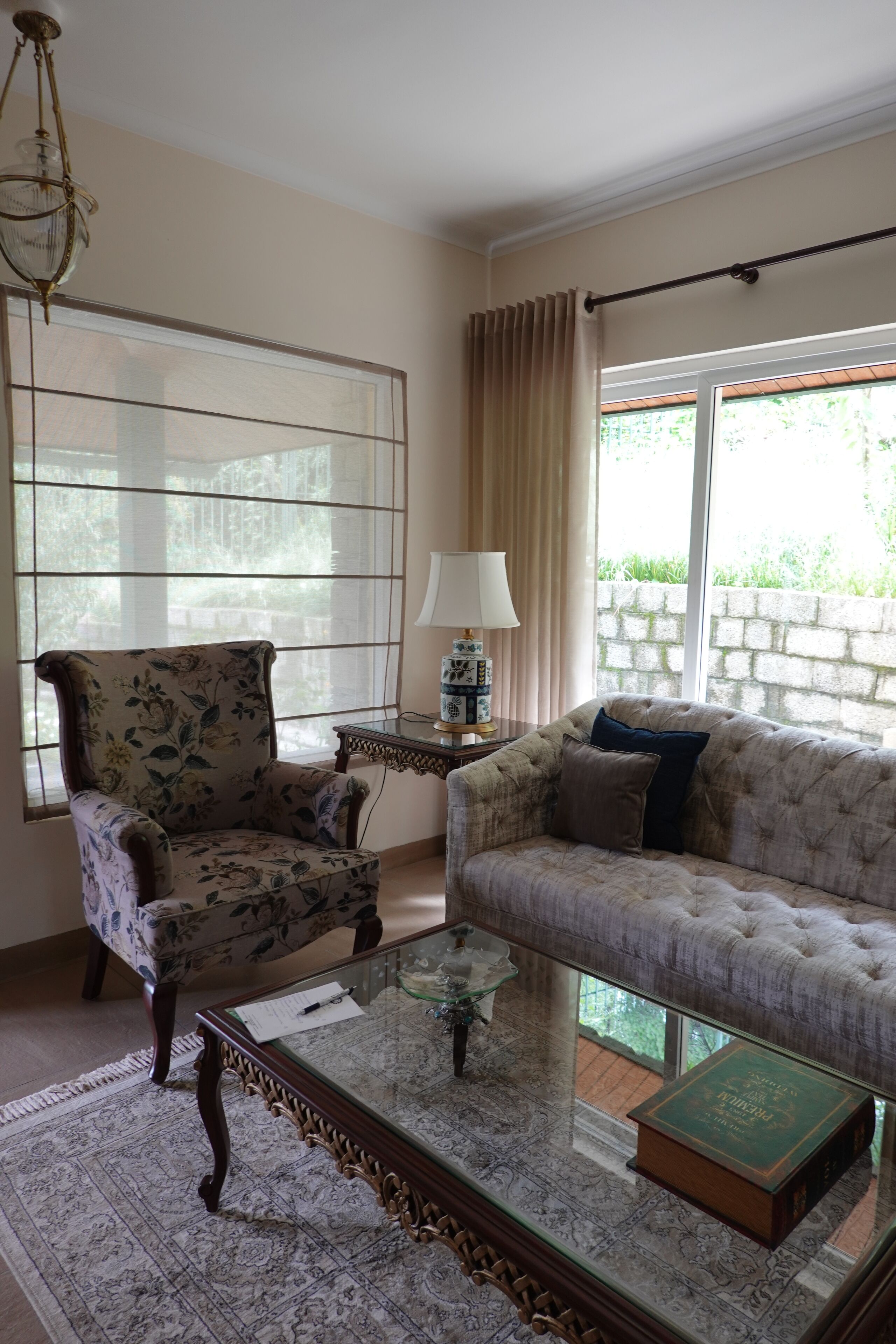 Una sala de estar con un sillón floral, un sofá capitonado, una mesa de centro de vidrio, una mesita de noche con una lámpara y vistas a través de una ventana con cortinas.