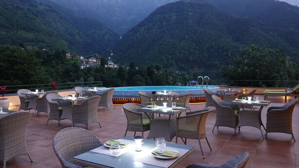 An outdoor dining area with wicker furniture and a pool in the background. - Araiya Palampur (Palampur)