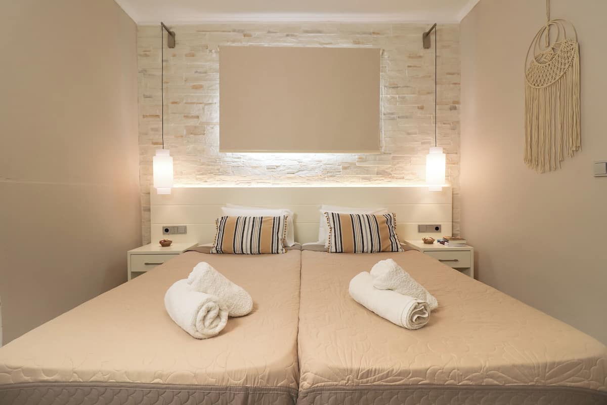 Een tweepersoonsbed met beige beddengoed, twee nachtkastjes met lampen, een aan de muur bevestigde televisie en een macramé wanddecoratie.
